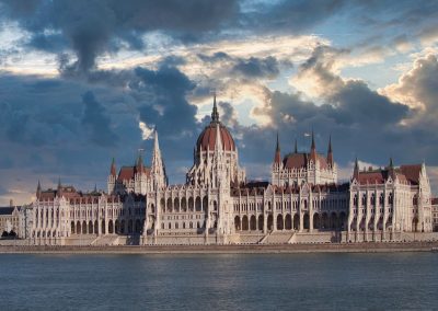 The Hungarian Parliament Building in Budapest stands majestically by the Danube River under a dramatic sky, as sunlight breaks through dark clouds, reflecting Hungary’s rich history.