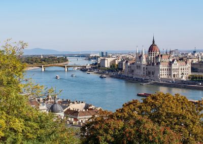 View of the Danube River in Budapest, Hungary, with boats on the water, the Hungarian Parliament Building on the right, a bridge crossing the river, and trees with autumn foliage painting a classic Hungary scene in the foreground.