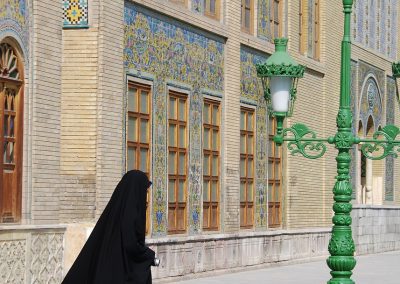 A person in a black chador walks past a decorative building with intricate tilework and wooden windows. A green ornate streetlamp stands in the foreground.