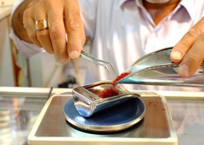 A person uses tweezers and a metal scoop to place red saffron threads onto a digital scale for weighing, wearing a white shirt and a ring on one hand.