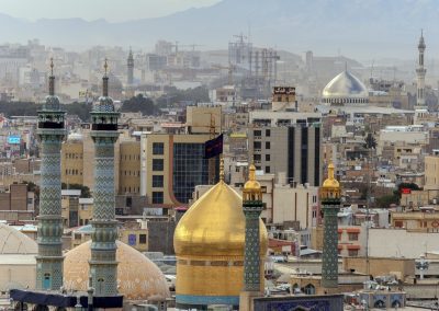 A cityscape of Qom, Iran, featuring ornate mosque minarets and a large golden dome in the foreground, with densely packed buildings and mountains visible in the hazy background.