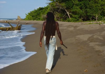 A person with long dreadlocks, wearing jeans and a sleeveless shirt, walks barefoot along a sandy Jamaican beach beside the sea, with green trees and driftwood in the background.