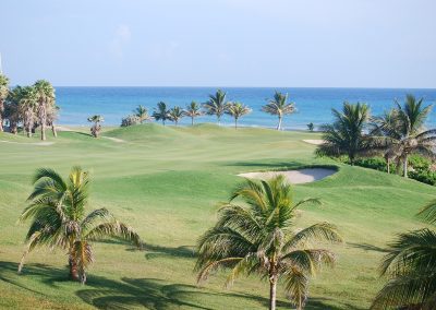 A coastal golf course in Jamaica with lush green fairways, palm trees, and a sand bunker, overlooking a calm blue sea under a clear sky.