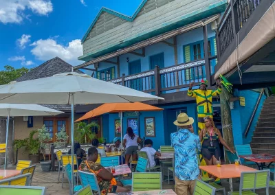 People sit at colourful outdoor tables under umbrellas at a vibrant café with blue walls. One person in a yellow and green outfit decorates the railing above, adding a lively touch of Jamaica as others relax and enjoy the sunny atmosphere.
