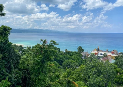 View of a lush green landscape in Jamaica with trees in the foreground, a small cluster of buildings to the right, and a calm blue sea stretching out to meet a partly cloudy sky in the background.