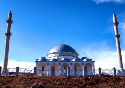 A white mosque with a blue dome and two tall minarets stands against a bright Kazakhstan sky, surrounded by a fence and dry brown ground in the foreground.