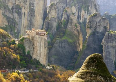 A monastery perched atop a tall, steep rock formation in Meteora, Greece, is surrounded by dramatic cliffs and lush greenery, with winding roads and trees below.