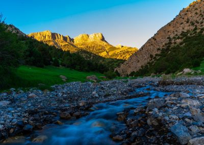 A rocky stream flows through a green valley surrounded by hills, with sunlight illuminating the distant mountain peaks under a clear blue sky.