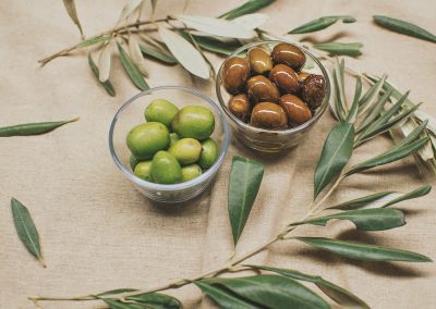 Two glass bowls filled with green and black olives are placed on a beige fabric, surrounded by olive branches and leaves, evoking the flavours and traditions of Greece.