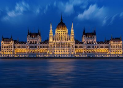 The illuminated Hungarian Parliament Building at night, reflected in the calm waters of the Danube River, captures the enchanting beauty of Hungary under a deep blue sky with scattered clouds.