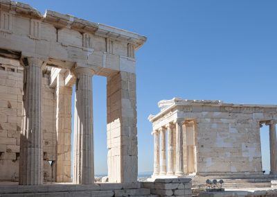 Ancient Greek temple ruins with tall stone columns and weathered marble, set against a clear blue sky in Greece, likely part of the Acropolis in Athens.