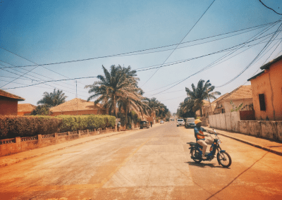 A person wearing a yellow helmet rides a motorbike across a sunny, palm-lined street in Guinea-Bissau, with houses and parked cars on both sides. The sky is clear and blue.