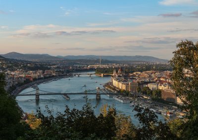 A panoramic view of Budapest, Hungary, with the Danube River running through the city, bridges connecting both sides, historic buildings, and hills in the background under a partly cloudy sky. Trees frame the foreground.