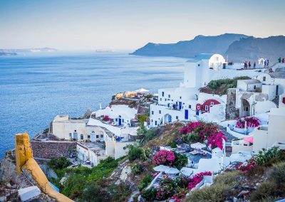 Whitewashed buildings with blue domes and pink flowers overlook a calm blue sea on a sunny day in Greece’s Santorini, with dramatic cliffs and distant islands visible across the water.