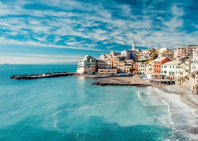 A vibrant coastal town in Greece with colourful buildings lining the shore, a small sandy beach, and turquoise sea under a partly cloudy blue sky. Waves gently lap the beach, and a church tower rises above the town.