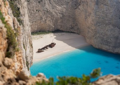 A secluded sandy beach in Greece, with turquoise water and steep cliffs, features a rusted shipwreck resting on the shore.