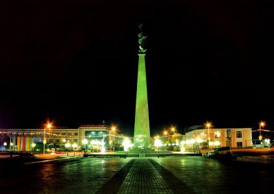 A tall illuminated obelisk monument with a statue on top stands in a wide plaza at night in Kazakhstan, surrounded by bright streetlights and buildings in the background.