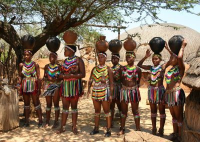 A group of people in colourful traditional Eswatini attire stand outdoors, balancing pots on their heads. They are surrounded by trees and thatched huts, with intricate beadwork and vibrant clothing on display.