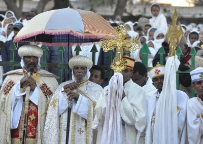 A group of Ethiopian Orthodox priests and followers dressed in ornate white and gold robes and headdresses participate in a religious ceremony, holding ornate crosses, with a large crowd in the background.