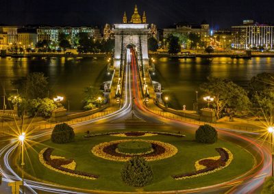 Night view of a brightly lit suspension bridge in Hungary with car light trails, crossing a wide river towards a cityscape of illuminated historic buildings and well-manicured flowerbeds in the foreground.