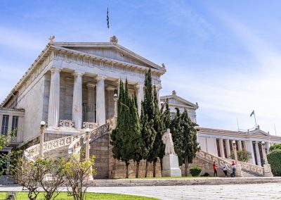 A neoclassical marble building in Greece with columns and grand staircases, surrounded by tall cypress trees and a statue in front, stands under a clear blue sky. People are walking near the entrance.