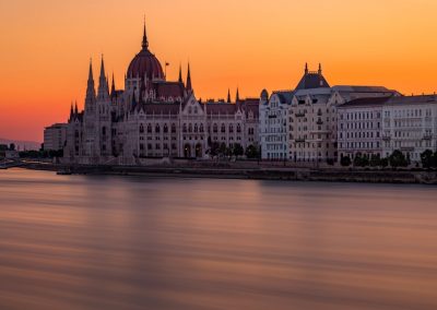 The Hungarian Parliament Building and nearby structures are silhouetted against an orange sunset sky, reflected in the calm waters of the Danube River in Budapest, Hungary.