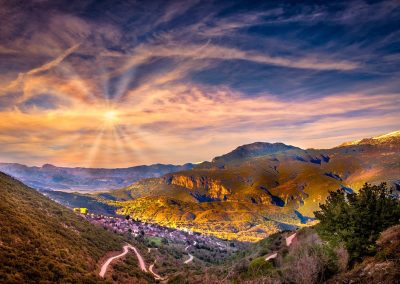 A vibrant mountain landscape in Greece at sunset, with sun rays shining through colourful clouds, winding roads, lush green valleys, and a small village nestled among the hills.