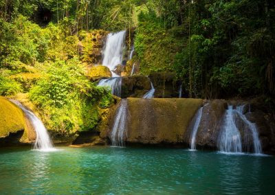 A lush, green tropical forest in Jamaica surrounds a tiered waterfall cascading over mossy rocks into a clear blue pool of water. Sunlight filters through the trees, highlighting the vibrant foliage.