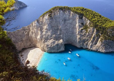 Aerial view of Navagio Beach in Greece, with striking white cliffs, turquoise water, a sandy cove dotted with people, a famous shipwreck on the shore, and several boats anchored in the bay.
