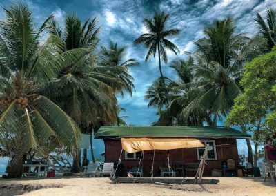 A small wooden cabin with a green roof sits on a sandy Micronesia beach, surrounded by tall palm trees under a dramatic, partly cloudy sky. A yellow canopy shades chairs and a swing in front of the cabin.