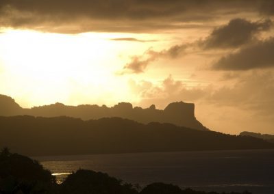 A golden sunset illuminates dramatic clouds and silhouettes of rugged hills and mountains beside a calm body of water in Micronesia.