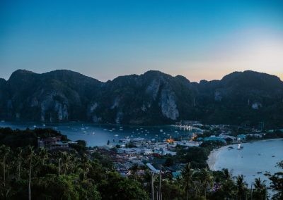 A coastal town nestled between lush green hills and the sea at dusk, with boats dotted on the water and buildings illuminated by warm lights.