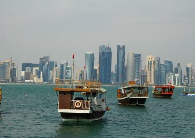 Traditional wooden boats float on calm water in Qatar, with a modern city skyline featuring tall skyscrapers in the background, under a hazy sky.