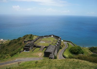 A historic stone fort sits on a grassy hill in Saint Kitts and Nevis, overlooking the blue ocean with winding roads leading up to it and lush green vegetation surrounding the area under a bright sky.