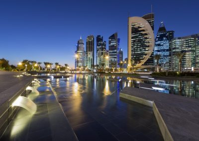 Modern cityscape at dusk in Qatar, with illuminated skyscrapers, unique curved buildings, palm trees, and a reflective water feature in the foreground against a deep blue sky.