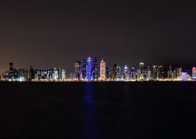 A night-time cityscape in Qatar featuring a brightly lit skyline with numerous high-rise buildings reflecting on dark, calm water under a clear night sky.