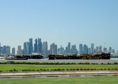 Traditional wooden boats are moored by the waterfront in Qatar, with a modern city skyline filled with tall skyscrapers rising in the background beneath a clear blue sky. Green grass and flowers add colour to the foreground.