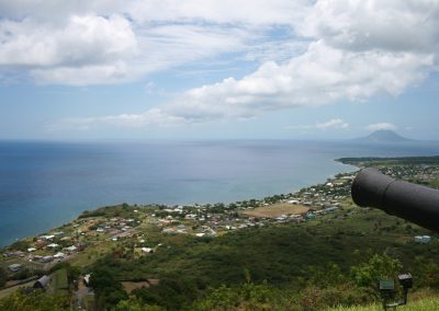 A coastal view from a hilltop in Saint Kitts and Nevis, showing a historic cannon overlooking a small seaside town, lush greenery, and calm blue sea with another island visible in the distance under a partly cloudy sky.