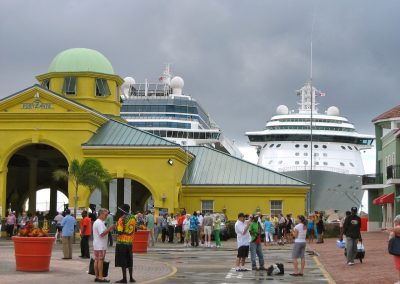 A bustling port scene in Saint Kitts and Nevis with a yellow building, crowds of people, and two large cruise ships docked under a cloudy sky. Palm trees and red planters decorate the vibrant area.