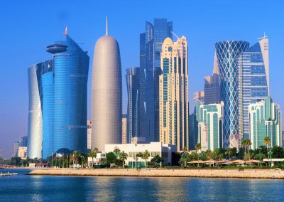 A waterfront view of Doha’s skyline in Qatar, with modern, uniquely shaped skyscrapers, glass façades, and palm trees under a clear blue sky, reflecting in the calm water.