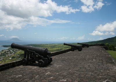 Three old cannons are positioned on a stone fort in Saint Kitts and Nevis, overlooking a lush, green landscape and coastline, with mountains and a partly cloudy sky in the background.