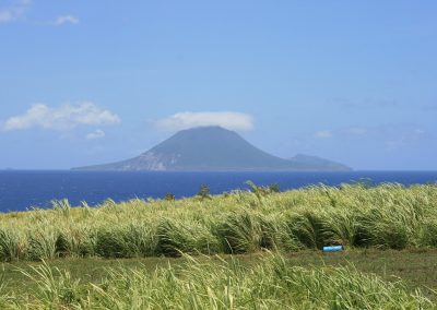 A grassy field in the foreground overlooks a calm blue sea, with the green, cone-shaped island of Saint Kitts and Nevis rising in the distance under a clear sky with scattered clouds.