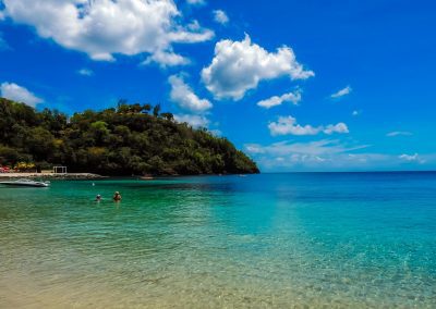 Clear turquoise water meets a sandy beach under a bright blue sky in Saint Vincent and the Grenadines, with a green, tree-covered hill in the background and a few people swimming near the shore.