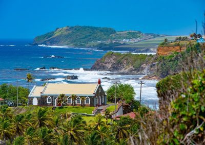 A stone church with a red roof sits on a grassy hill overlooking the sea in Saint Vincent and the Grenadines, surrounded by palm trees and lush cliffs under a bright blue sky. Waves crash against the rocky coastline in the background.