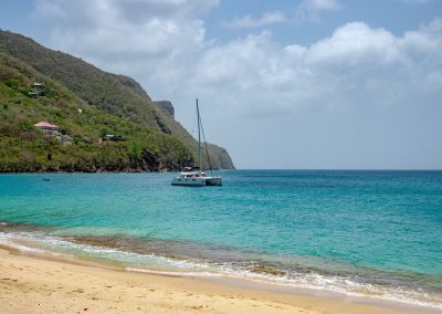A white catamaran floats on clear turquoise water near a sandy beach in Saint Vincent and the Grenadines, framed by green hills and a few houses in the background under a partly cloudy sky.