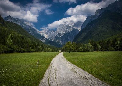 A winding tarmac path leads through a green field in Slovenia towards distant, forested mountains under a partly cloudy sky, with tall peaks rising dramatically in the background.