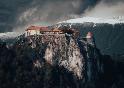A stone castle with red roofs sits atop a steep, rocky cliff surrounded by dense forest in Slovenia, with snow-capped mountains and dramatic, cloudy skies in the background.