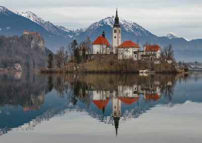 A small island with a white church and red roofs sits on a calm Slovenian lake, reflecting perfectly in the water. Snow-capped mountains and a cloudy sky are in the background, creating a peaceful, scenic landscape.
