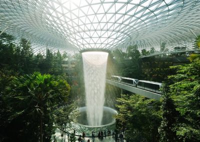 A large indoor waterfall cascades from a glass dome ceiling surrounded by lush greenery and palm trees in Singapore. A monorail train glides along an elevated track in the background, as people gather around the breathtaking waterfall below.