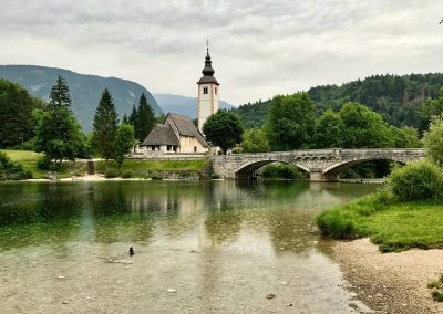 A stone bridge crosses a calm lake in Slovenia near a small church with a tall spire, surrounded by lush green trees and mountains under a cloudy sky. A duck swims near the shore in the foreground.